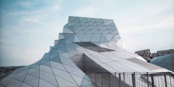 Futuristic building with geometric, angular design featuring a facade of triangular patterns and cutting-edge metal roofing. The structure reflects light under a blue sky with scattered clouds, overlooking the distant cityscape. A top-notch roofing contractor ensured its impeccable finish.
