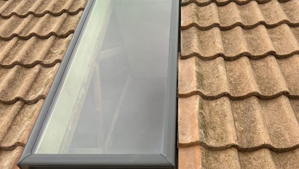 A skylight window is installed on a red-tiled roof, showcasing expert roofing craftsmanship. The rectangular window with a black frame sits amid a slightly uneven pattern of clay tiles, while the overcast sky looms above.