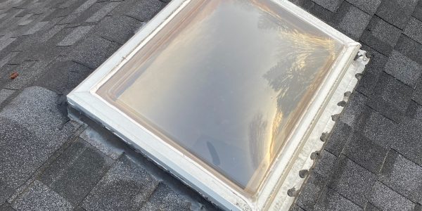 A rectangular skylight with a semi-transparent dome is installed on a dark shingle roof, recently serviced by a roofing company. The roof features a ventilation turbine nearby, and autumn leaves are visible on the ground below in the background.