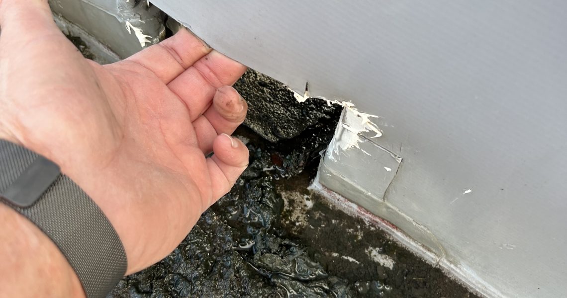 A persons hand is pulling back a torn section of white material on a wall, revealing damage underneath. The ground is covered in black, sticky dirt and debris. A roofing contractors watch glints as they assess the necessary repairs.