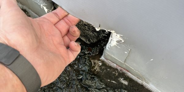 A persons hand is pulling back a torn section of white material on a wall, revealing damage underneath. The ground is covered in black, sticky dirt and debris. A roofing contractors watch glints as they assess the necessary repairs.