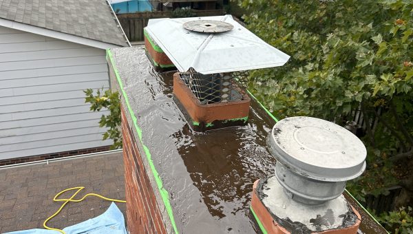 A brick chimney with two metal caps is shown from above, surrounded by fresh cement and green tape outlining the edges. Nearby, a blue tarp partially covers roofing materials on the roof. In the background, trees frame a backyard complete with a fence and pool.