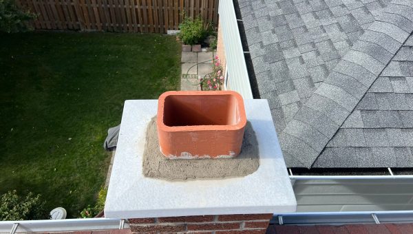A red brick chimney topped with a square, orange flue enveloped in gray mortar stands on a roof with gray shingles. In the background, a fence and green backyard can be seen—a testament to quality craftsmanship by your local roofing company.