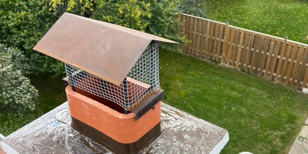 A birds-eye view of a backyard reveals a brick chimney, likely maintained by a roofing contractor, in the foreground. The chimney has a rectangular metal and mesh cover. The yard features a lawn, wooden fence, trees, patio with chairs, and a barbecue grill under a cover.