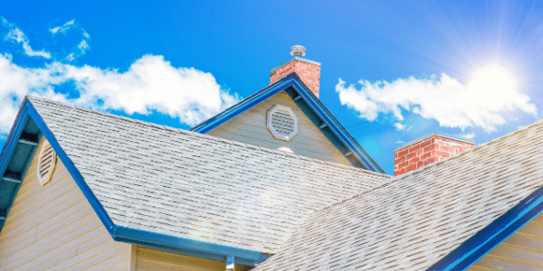 Sunny view of a house roof with blue trim, expertly crafted by a roofing contractor, chimney, and vent set against a bright blue sky with scattered clouds and a radiant sun.