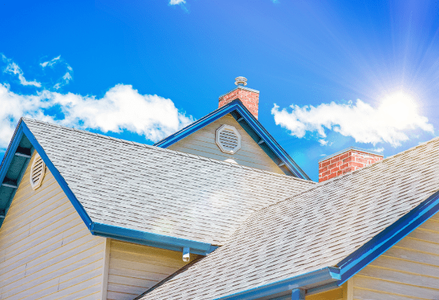 Sunny view of a house roof with blue trim, expertly crafted by a roofing contractor, chimney, and vent set against a bright blue sky with scattered clouds and a radiant sun.