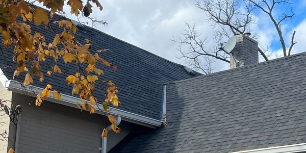 A house with a gabled roof covered in gray shingles, expertly maintained by a roofing contractor. A chimney and satellite dish stand visible. Autumn leaves from a nearby tree frame the top left corner. The sky is partly cloudy and blue.