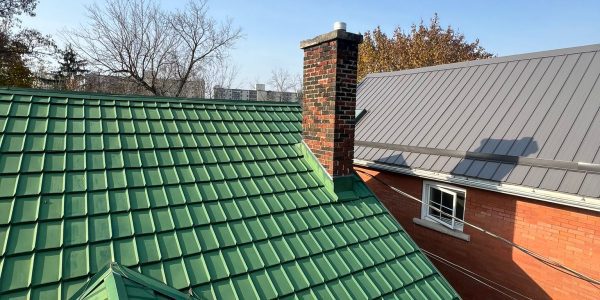 View of a green metal tiled roof showcasing quality metal roofing with a red brick chimney, beside a red brick building with a gray metal roof, under a clear blue sky and bare trees in the background.
