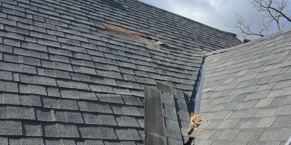 A roof with missing shingles reveals underlying wood, suggesting a call to a roofing company. The dark gray shingles contrast with the bright blue sky and scattered clouds. A ladder is partially visible on the right, and a leafless tree branch can be seen in the background.