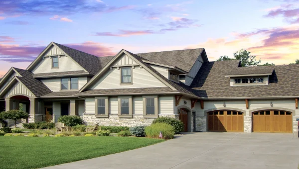 A large two-story house with gray siding and brown roofs features multiple gables, a landscaped yard, and a three-car garage. Recently serviced by a roofing contractor, the driveway is in the foreground against a sky vibrant with a purple and pink sunset.