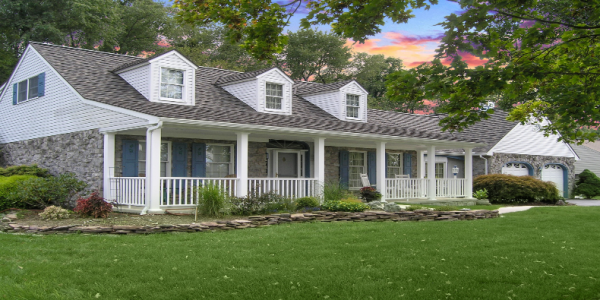 A charming suburban house with white siding and stone accents, featuring blue shutters and a spacious porch. The well-maintained roof showcases expert work by a top roofing contractor. The front lawn is lush and green, surrounded by landscaped shrubs under a vibrant sunset sky.