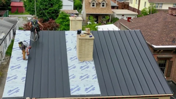 Two workers, acting as roofing contractors, are installing dark-colored metal roofing panels on a house. The middle section is lined with insulation sheets labeled Union Corrugating. Surrounding the house are other residential buildings and greenery.