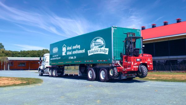 A large truck with a trailer showcasing the Westfield Bridge logo for roofing and construction services, including skylight repair. A red forklift is attached to the back. The truck is parked on a gravel path beside a red and black industrial building, under a clear blue sky.