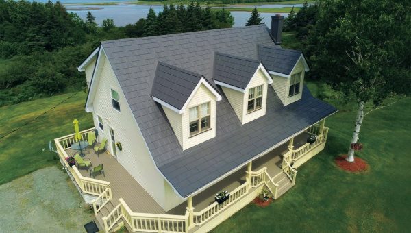 A large white house with a grey roof, featuring dormer and skylight windows along a wraparound porch adorned with outdoor furniture. Set in a lush green landscape near water, its surrounded by trees under a partly cloudy sky, offering the perfect setting for any necessary skylight repair.