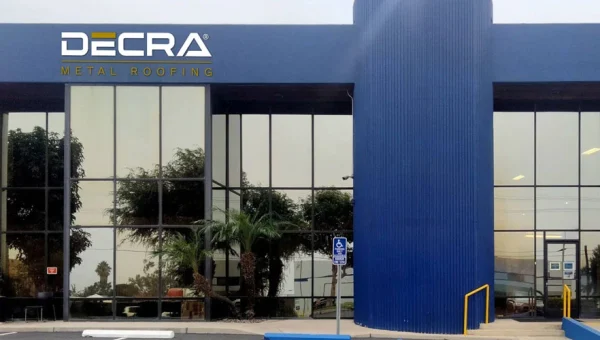Modern commercial building with a blue facade and large glass windows, showcasing the DECRA Metal Roofing logo on the upper left. As a prominent roofing company, its reflective surfaces reveal palm trees outside. Accessible parking and entrance are visible in the foreground.