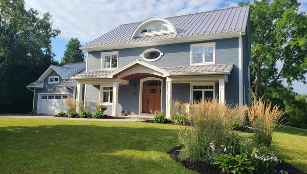 A two-story blue house with a metal roof, expertly installed by a roofing contractor, is surrounded by a lush lawn and landscaped plants. It features a large wooden front door, white trim, and an attached garage. The sky is partly cloudy, with trees beautifully framing the scene.