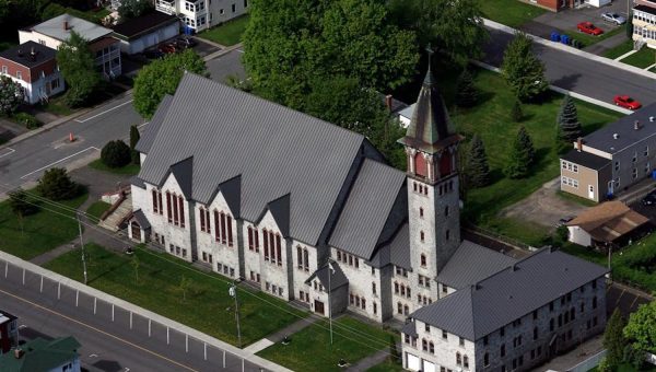 Aerial view of a large, stone church with a gray roof and tall clock tower. The scene captures the essence of suburbia, with residential buildings, trees, and roads encircling it. The sturdy roofing frames the skyline beautifully amid whispers of skylight repair tales in neighboring homes.