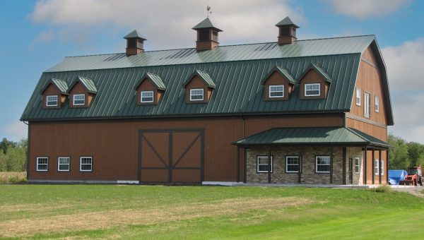 A large brown barn with a green metal roof, possibly crafted by a skilled roofing contractor, stands on a well-kept green lawn. It features multiple windows and dormers. The sky is partly cloudy, and a vehicle is partially visible on the right.