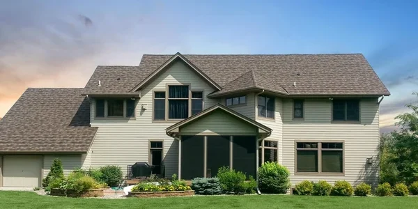 A large two-story house with beige siding and a dark brown roof, expertly maintained by a top roofing contractor. It features multiple windows and is set in a spacious, well-manicured yard with green grass and some shrubs, under a clear blue sky.