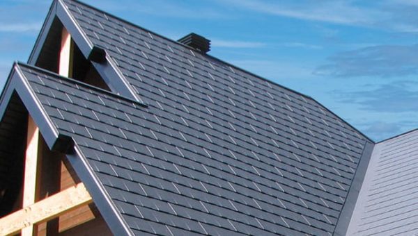 A modern house roof with dark gray, sleek shingles against a clear blue sky features multiple gables and a chimney. The clean, organized pattern showcases neat architectural design, complemented by a recent skylight replacement for enhanced daylighting.