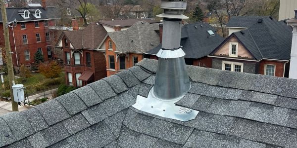 From a roof with gray shingles, expertly crafted by a skilled roofing contractor, see the silver chimney pipe standing tall. Below lies a neighborhood of brick houses, overshadowed by a towering building. The sky is overcast with clouds.