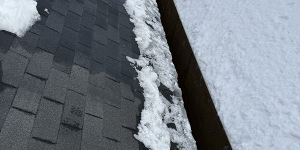 A roofing contractor in an orange vest is diligently shoveling snow on a shingled roof. Part of the roof is clear, with remaining snow piled along the edge, while the adjacent roof remains snow-covered. Its a snowy winter day.