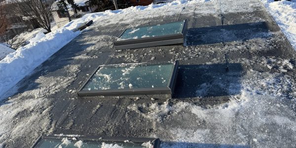 A flat rooftop with three rectangular skylights is covered in snow and patches of ice, hinting at potential roof repair needs. Surrounding buildings and trees stand under a clear blue sky.