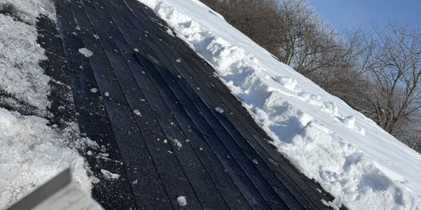 Snow partially covers a rooftop, revealing dark shingles that may need roof repair. A metal edge is visible in the foreground, while trees and a tall building rise in the distance against a clear blue sky.