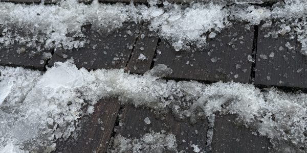 Snow and ice cover a shingled roof, with patches of roof visible between the snowy areas. The mixture of snow and ice creates a textured surface across the shingles, emphasizing the need for timely roof repair to prevent further weather damage.