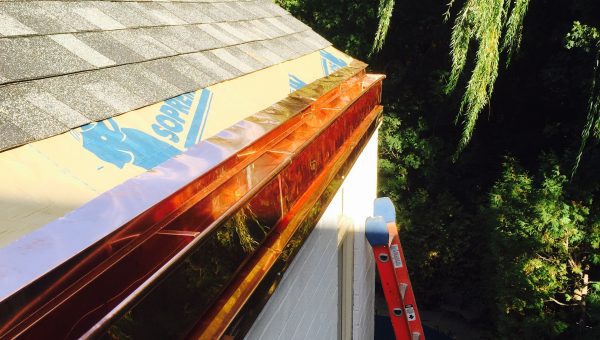 A roof under construction features new copper gutters along the edge, with a red ladder leaning nearby as green trees surround the scene. The sky is clear, revealing partially visible shingles and a chimney repair in progress.