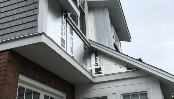 A house exterior with a mix of brick and white siding features partially installed insulation material on the upper part. Multiple windows with white frames accent the gables under an overcast sky, while a roofing contractor completes skylight repair work.