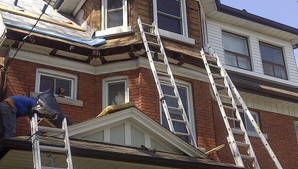 Workers are renovating the exterior of a multi-story brick house. Ladders are propped against the walls, and a portion of the siding is removed on the upper floor. A roofing contractor is visible near a window, engaged in repairs alongside chimney work.