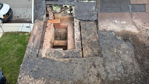 A damaged roof with missing shingles and exposed bricks surrounds a large rectangular hole near the chimney. The surrounding shingles and roofing material appear worn, with a street and lawn visible in the background.