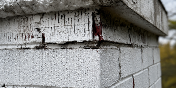 Close-up of a white brick corner with peeling paint and rough, weathered texture. Some red underlayers are visible through cracks. The sky and trees appear blurred in the background.